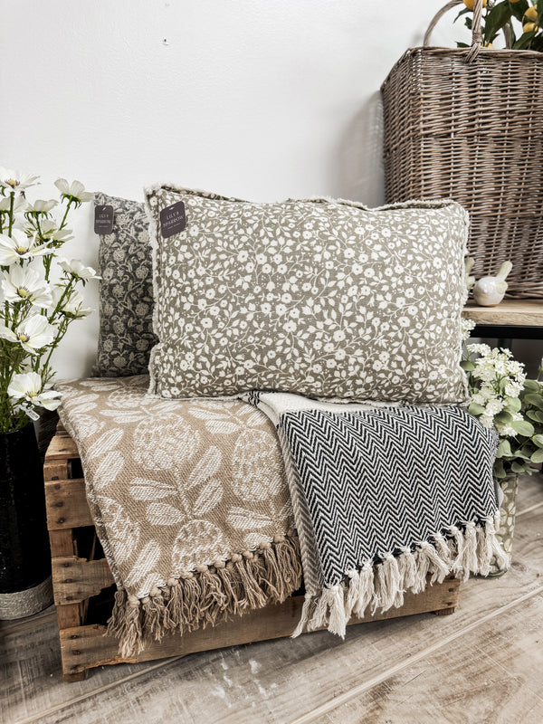 Decorative pillows and blankets on a wooden bench with a wicker basket and flowers in the background.